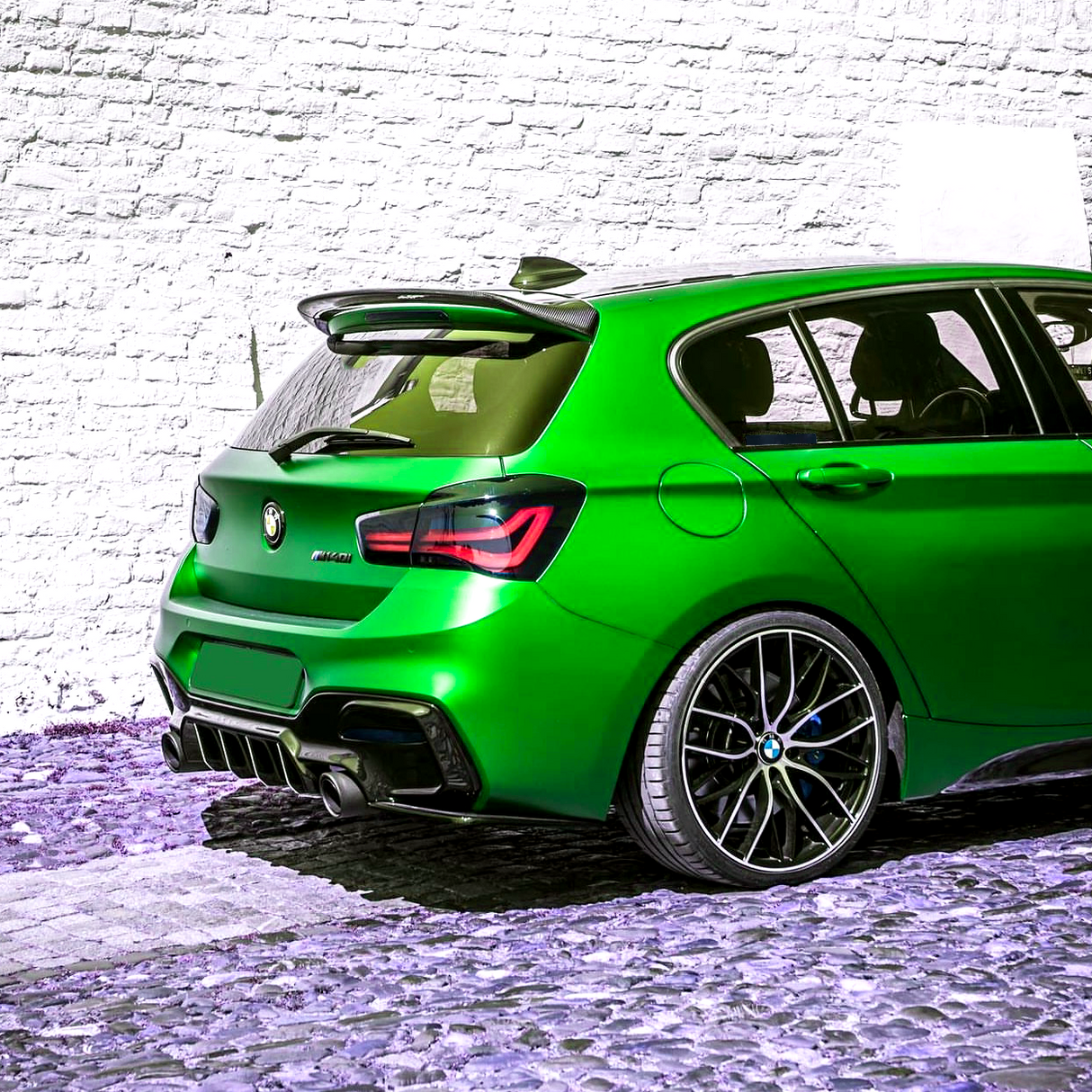 A bright green hatchback car is parked on a cobblestone path, situated against a textured white brick wall.