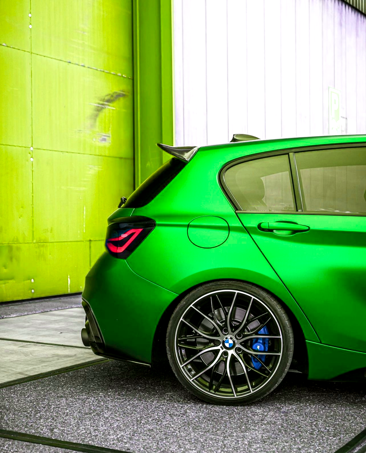 Green sports car parked near a yellow and white wall. The image focuses on the rear wheel, which features a blue brake caliper and distinctive alloy design.