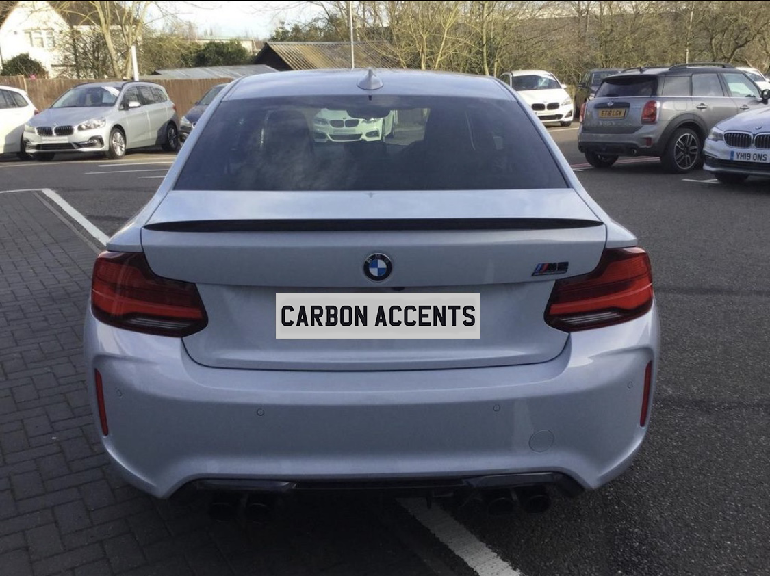 A silver BMW car is parked, displaying the text "CARBON ACCENTS" on its rear plate. It's situated in a parking lot with various other vehicles nearby.