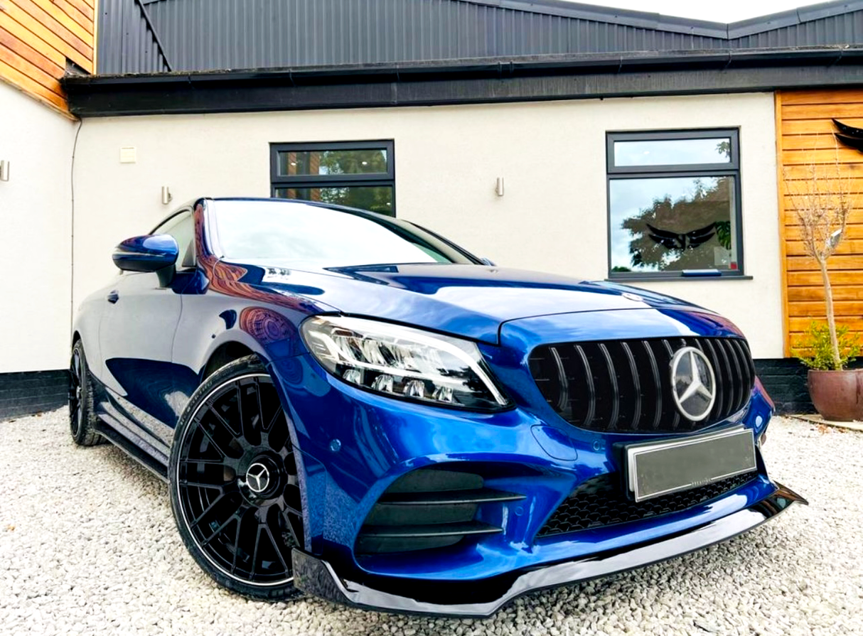 Blue Mercedes car parked on gravel, showcasing black wheels and grille. The background features a modern house with white walls, wooden paneling, and large windows.