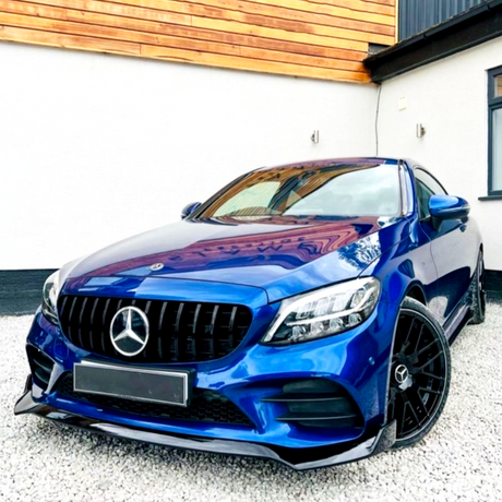 A blue Mercedes-Benz car is parked on a gravel driveway, surrounded by a white house with wooden panel accents. The vehicle features black wheels and a prominent front grille.