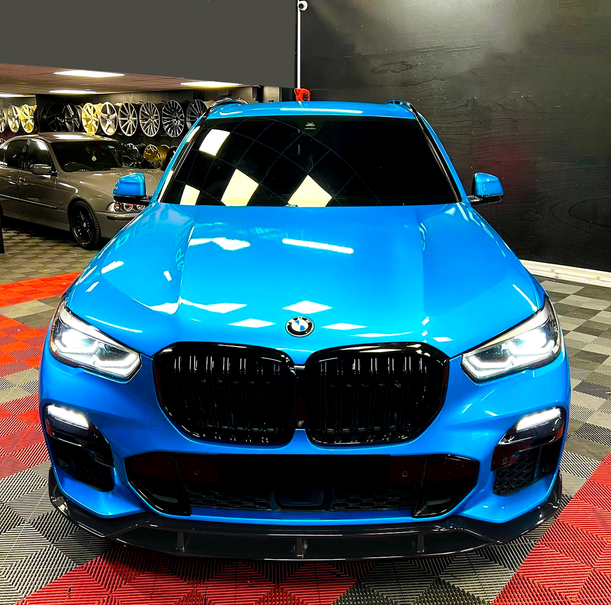 A blue BMW SUV is parked with headlights on in a showroom, surrounded by various wheels displayed on the wall and a checkered floor pattern.