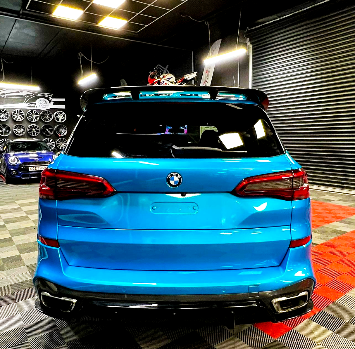 A bright blue BMW SUV is stationary in a garage with shelves of wheels and vehicles, like a blue sports car. The floor features checkered patterns, and lighting is overhead.
