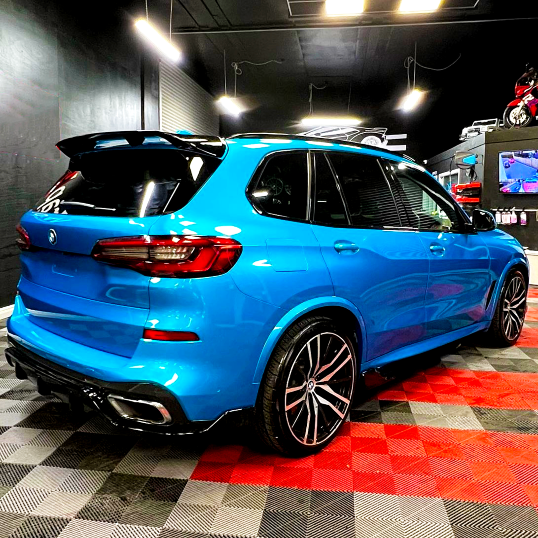 A blue SUV is parked on a red and gray checkered floor in a well-lit garage. The garage has black walls, overhead lights, and a television displaying images.