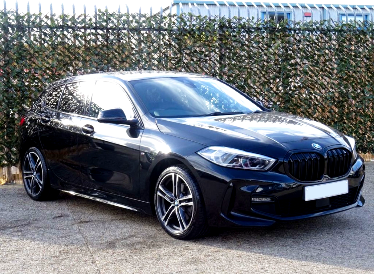 A sleek black car parked on pavement, reflecting sunlight. It's positioned in front of a fence covered with green camouflage netting, with a building partly visible behind.