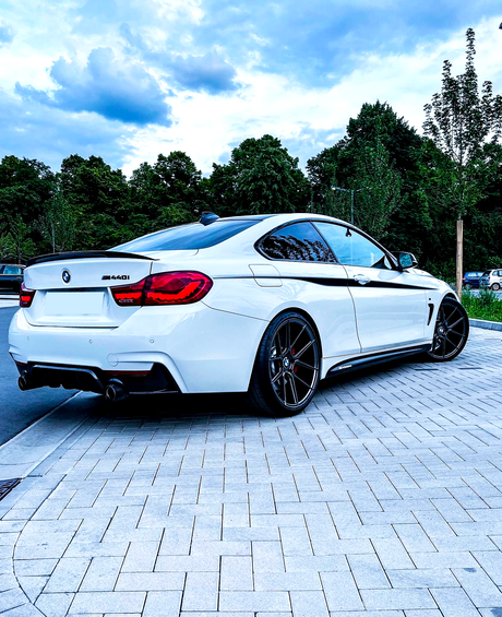 A white BMW 440i is parked on a paved street, against a backdrop of lush green trees and a cloudy sky.