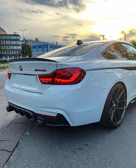 A white BMW M440i is parked on a city street at sunset, featuring illuminated red taillights and dual exhaust pipes, adjacent to modern buildings.