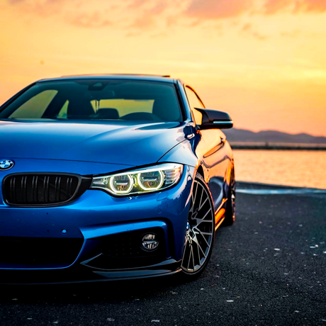 A blue car is parked by a waterfront at sunset, with its headlights on and mountains in the background.
