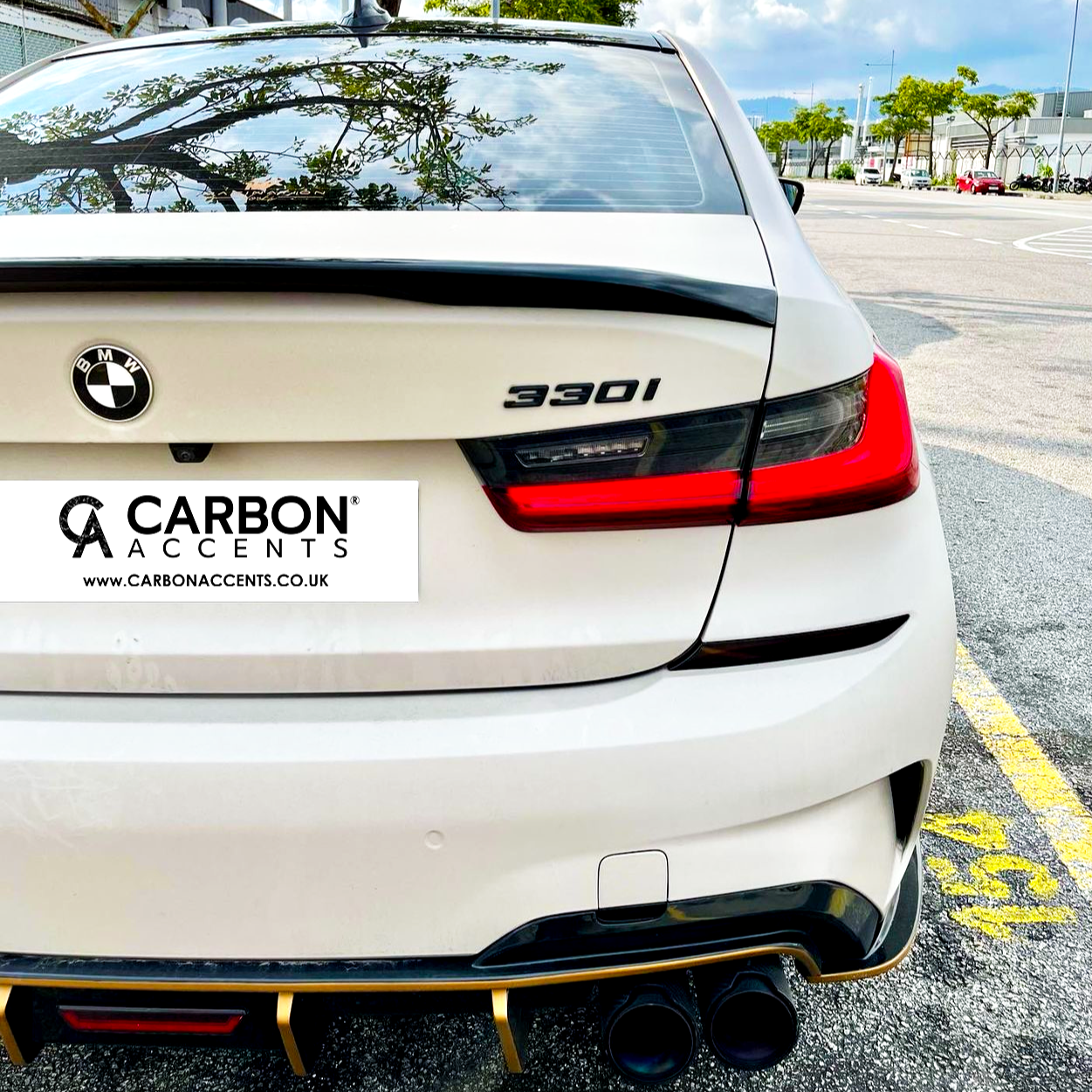 BMW car rear showcasing "330i" badge and dual exhausts; parked in a lot with trees and buildings visible. Text: “CARBON ACCENTS” and “www.CARBONACCENTS.CO.UK”.