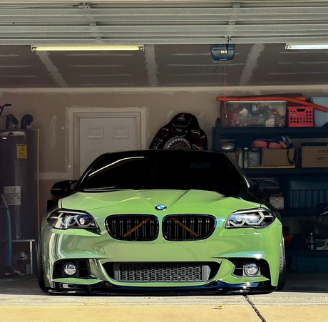A sleek green car is parked in a cluttered garage, surrounded by shelves filled with boxes and various household items.