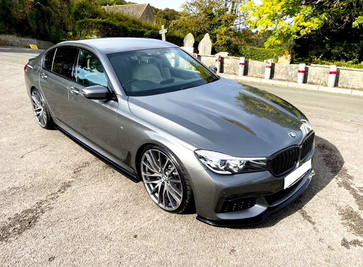 A sleek, grey sedan is parked on a quiet road beside stone markers and lush greenery, under a clear sky.