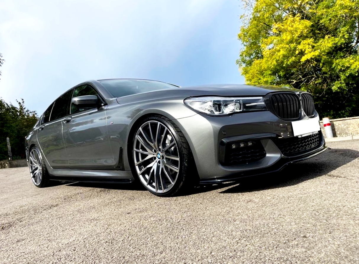 A sleek grey car parked on a paved surface with large alloy wheels, surrounded by greenery on a clear day.