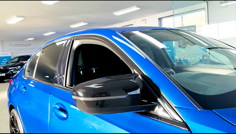 A blue car parked indoors, with its window slightly open and a visible side mirror reflecting the ceiling lights. The environment is a well-lit showroom with visible signage.