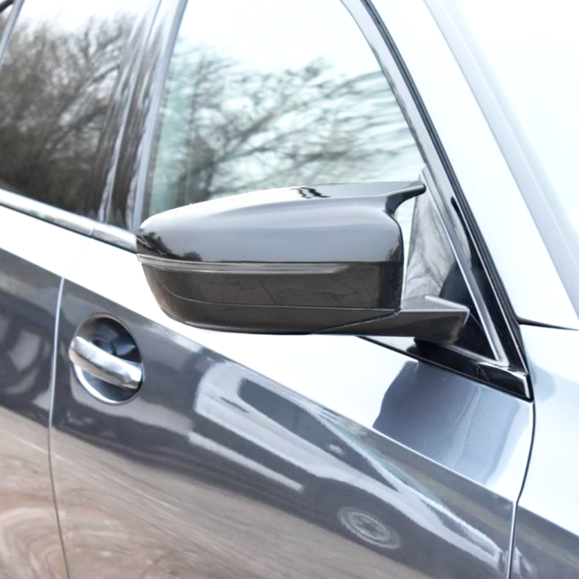 A car side mirror extends from a partially visible vehicle, reflecting trees. The vehicle is metallic gray, showcasing a sleek, modern design with a visible door and handle.
