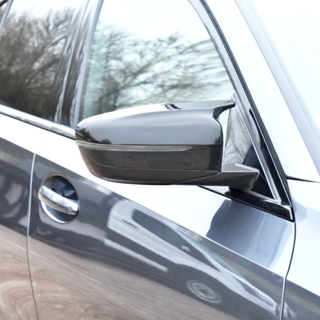 A car side mirror extends from a partially visible vehicle, reflecting trees. The vehicle is metallic gray, showcasing a sleek, modern design with a visible door and handle.