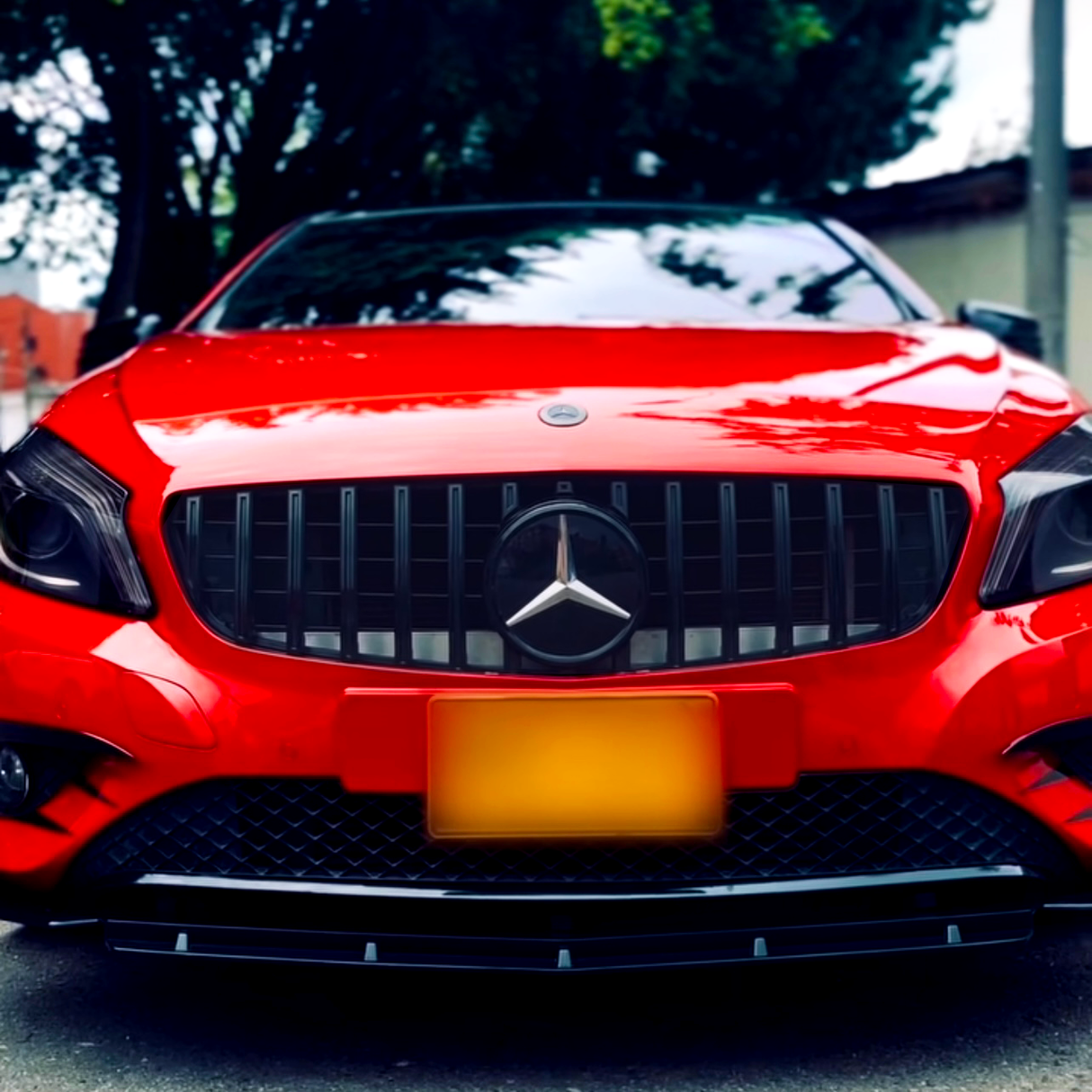 Red car is parked, facing forward. Its grille with a central emblem is prominent, surrounded by sleek headlights. Background shows trees and a building, creating an outdoor setting.