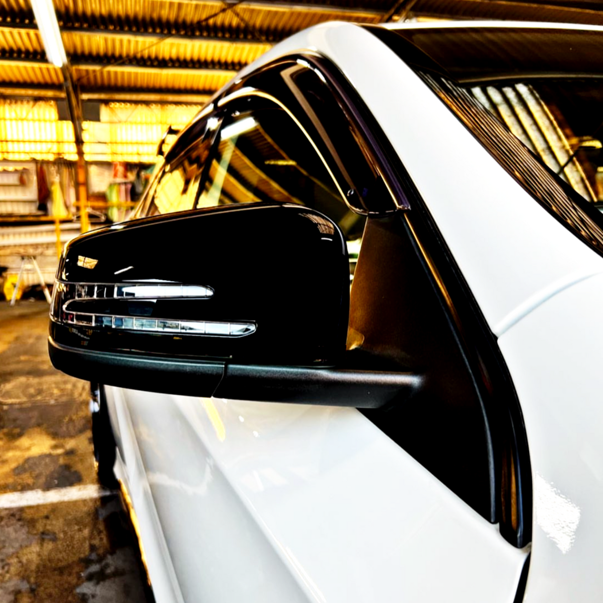 A black side mirror reflects on a white car, parked in a spacious, industrial garage with a corrugated metal roof and fluorescent lighting.