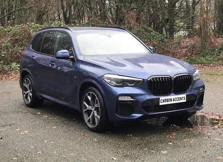 A blue SUV with prominent wheel rims is parked on a wet, leaf-strewn road. The license plate reads "CARBON ACCENTS." In the background, bare trees and shrubs create a natural setting.