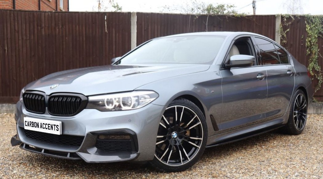 A silver car with sleek design and black wheels is parked on a gravel surface, with a wooden fence and brick structure in the background. Text on the license plate reads "CARBON ACCENTS."
