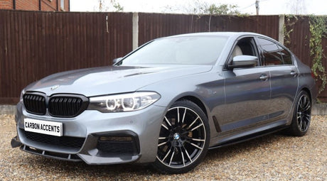 A silver car with sleek design and black wheels is parked on a gravel surface, with a wooden fence and brick structure in the background. Text on the license plate reads "CARBON ACCENTS."