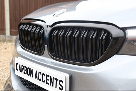 Car grille featuring vertical black slats prominently displayed on a front bumper. Below, a license plate reads "CARBON ACCENTS." The vehicle is parked on a gravel surface.