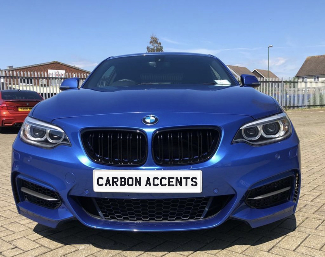 A blue BMW car is parked on a paved area under a clear sky, displaying a license plate with the text "CARBON ACCENTS." Other cars and buildings are in the background.