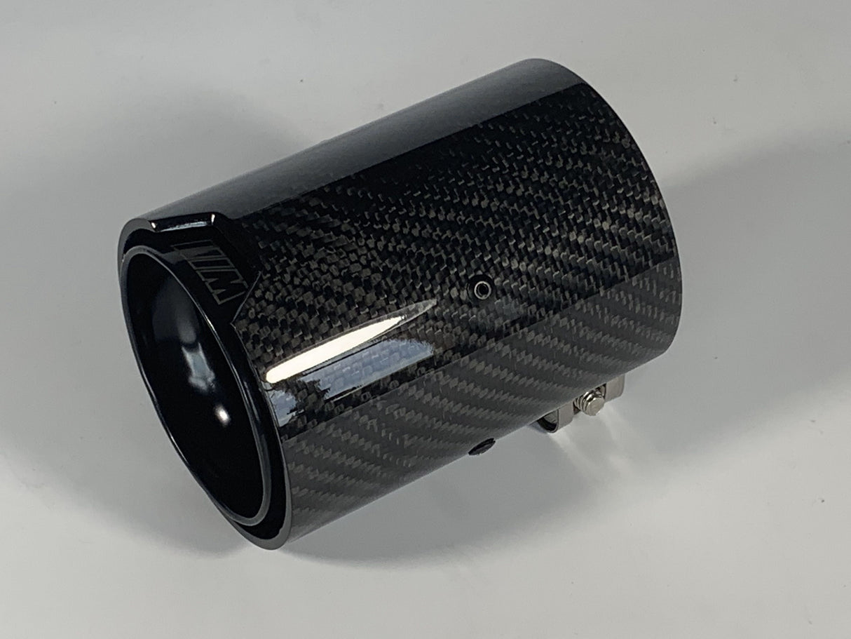 A cylindrical carbon fiber exhaust tip displays reflective surface details, resting on a solid white background. It features a small logo with stylized letters "MW" on the side.