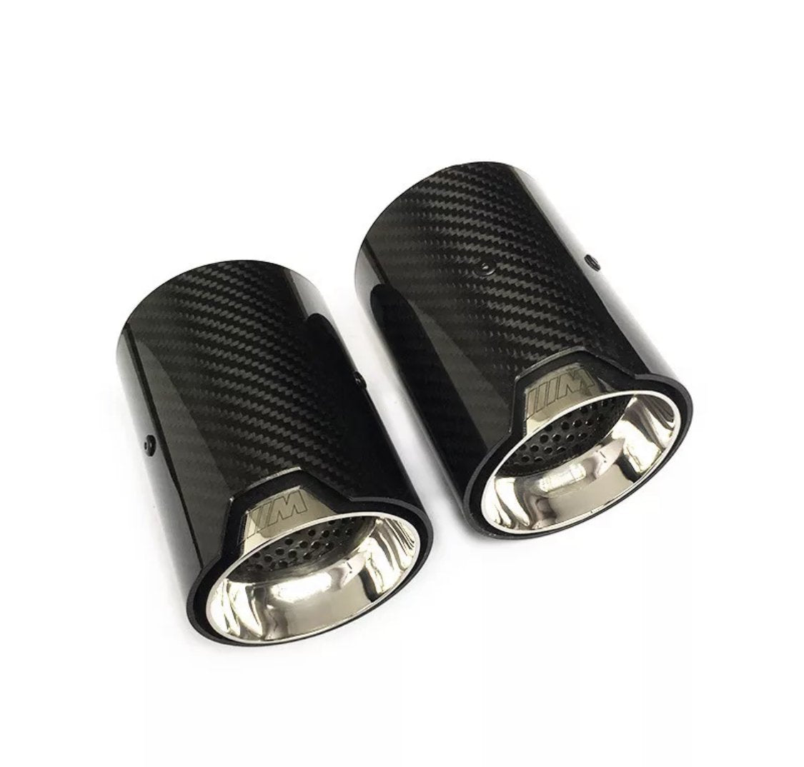 Two carbon fiber exhaust tips rest side by side. The shiny, metallic interiors reflect light. "M" emblem is visible near the opening. Placed on a plain white background.