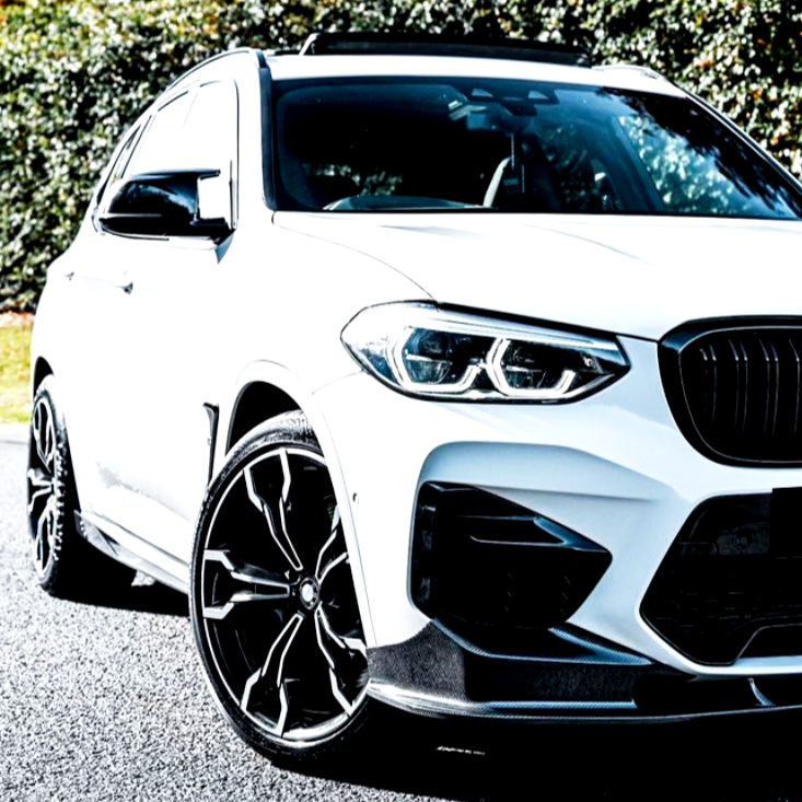 A white SUV with sleek black alloy wheels rests on a paved surface, surrounded by greenery. The vehicle's front grille and headlights are prominently displayed.