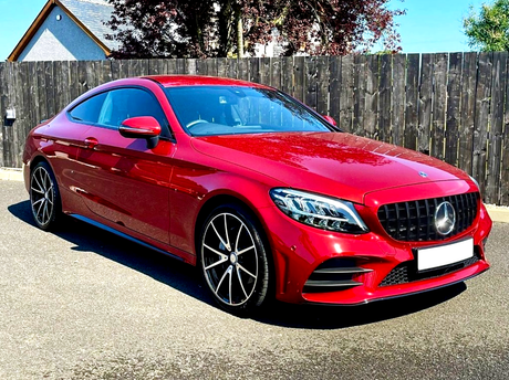 A red sports car is parked on a paved driveway. It features a sleek design with shiny black wheels. The wooden fence and a house with trees form the background.