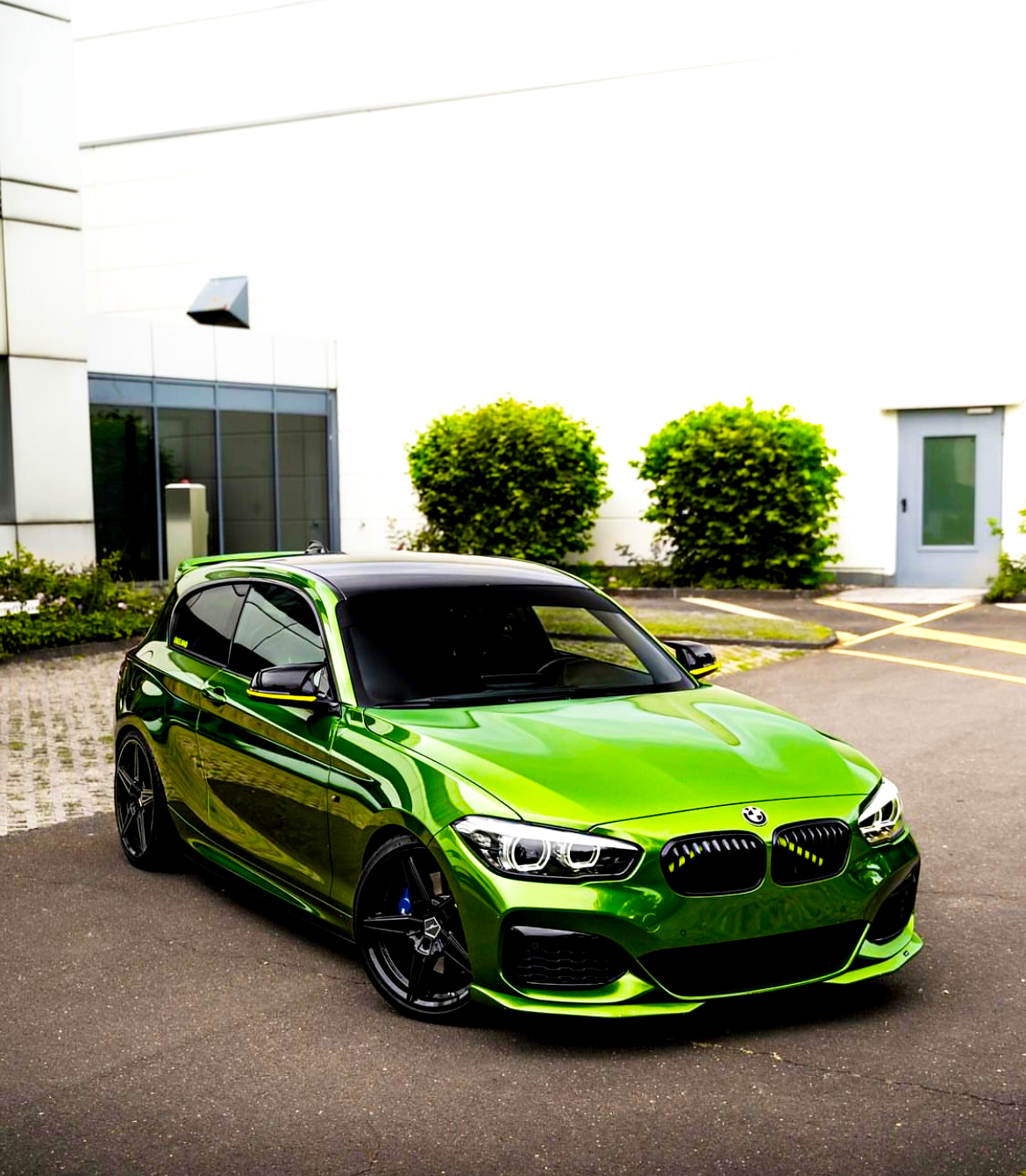 A glossy green car is parked on a paved driveway. Surrounding it are a white building and green shrubs, with a modern entrance visible in the background.