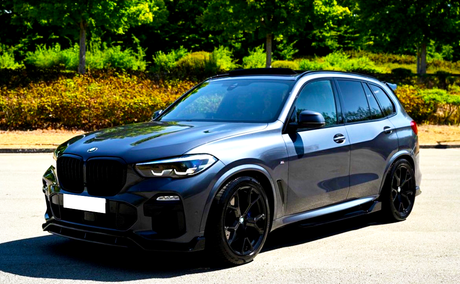 A sleek, dark-colored SUV is parked on a sunlit road, surrounded by lush green trees and bushes in the background.