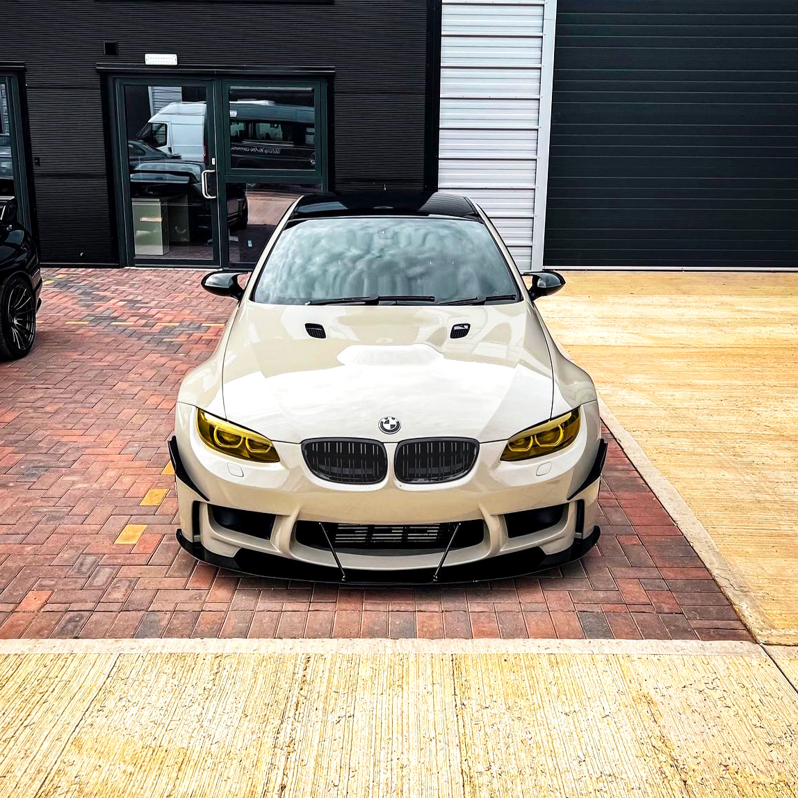 A white BMW car sits parked on a brick driveway in front of a modern building, featuring yellow-tinted headlights and a sleek, aggressive front design.