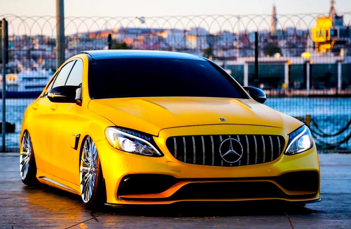 A yellow Mercedes-Benz car is parked on a paved surface beside a waterfront, featuring a distinctive grille and large chrome wheels, with a cityscape visible in the background.