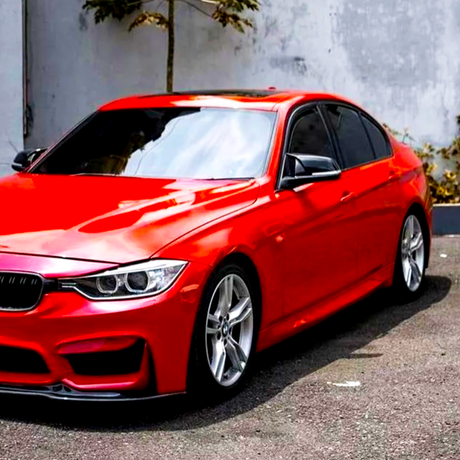 A red sedan parked on a concrete surface, with a background of a gray wall and a small tree. The car has tinted windows and silver alloy wheels.