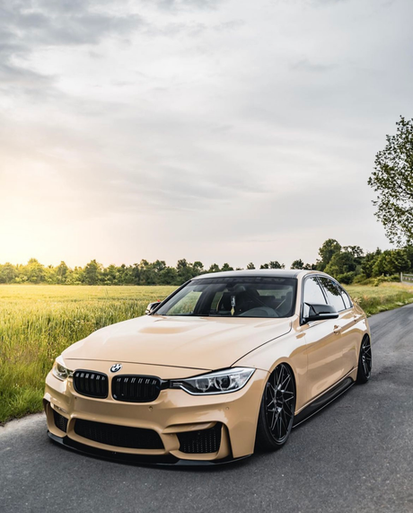 A beige sports car is parked on a paved road, surrounded by green fields and trees, under a cloudy sky, with sunlight casting a warm glow.
