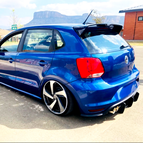 A blue hatchback car with custom rims and spoiler is parked on a paved ground. In the background, there's a brick building and a mountain under a partly cloudy sky.