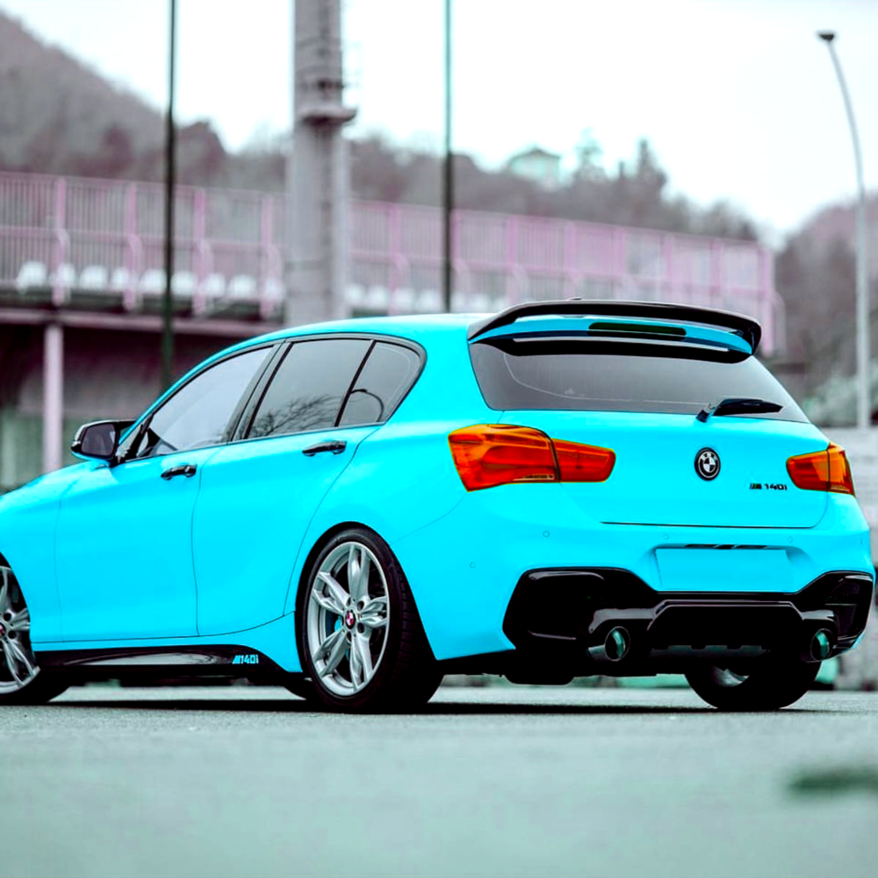 Blue car parked outdoors, showcasing its rear on gray pavement. Red taillights contrast with the bright color. In the background, a fenced area and blurred hilly terrain are visible.