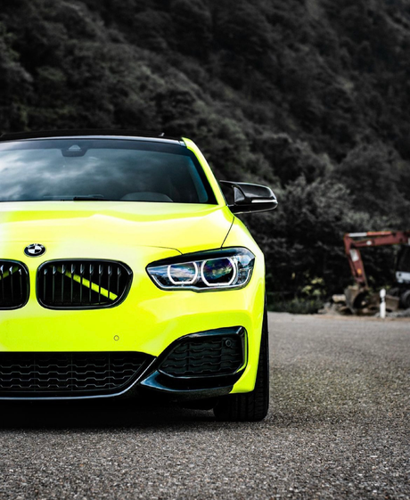 A bright yellow BMW car stands parked on a road, its sleek design contrasting with a forested hillside in the background. The environment appears rural with scattered machinery visible.