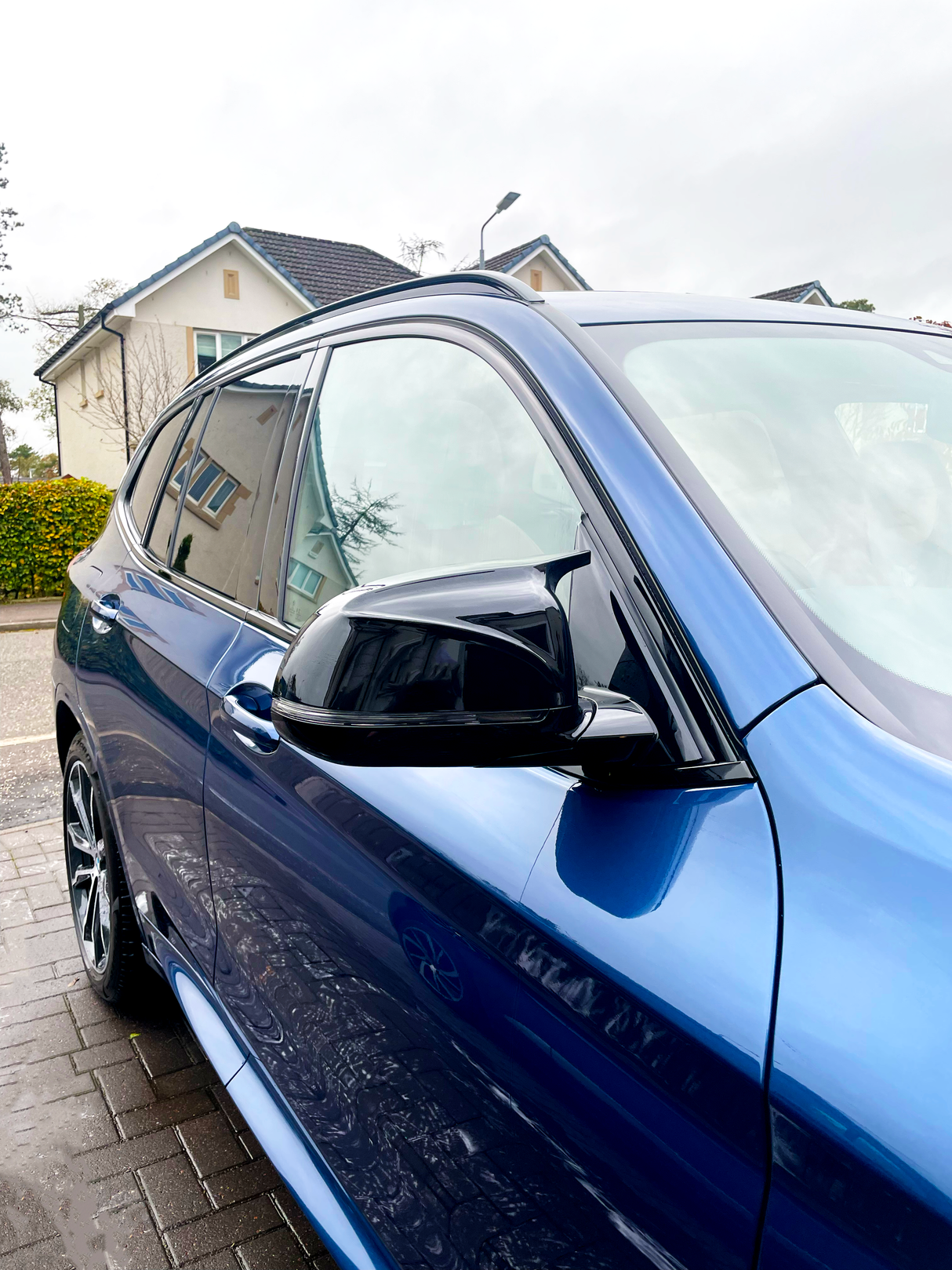 A blue car is parked on a wet driveway, showing a side mirror and window. It reflects nearby houses and a cloudy sky in a suburban neighborhood.