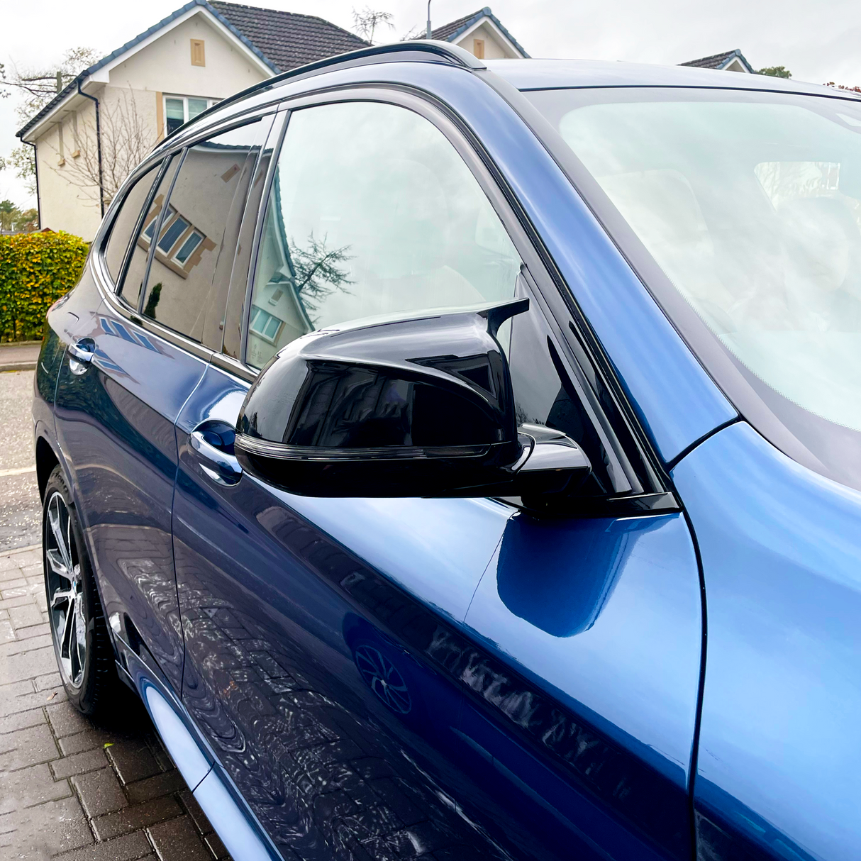 A blue car is parked on a driveway. The shiny side mirror and windows reflect neighboring houses and trees, creating a vivid, mirrored image of the suburban environment.