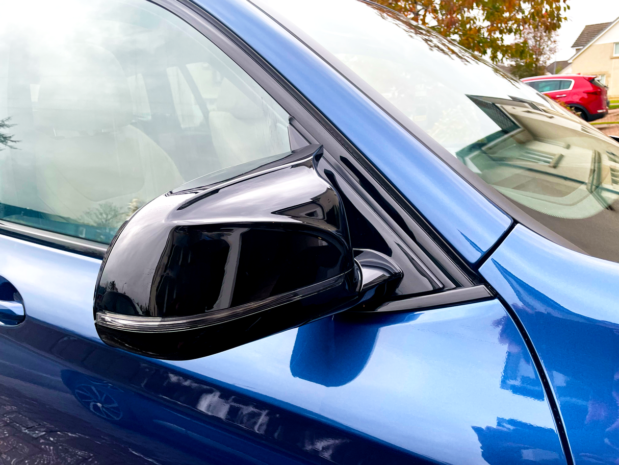 A shiny black side mirror is attached to a blue car, shown from the front passenger side. The background includes suburban houses and parked vehicles, suggesting a residential area.