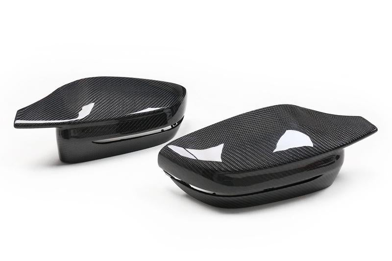 Two carbon fiber side mirror covers rest on a white background, showcasing a sleek, glossy finish with a subtle, woven pattern and reflection details.