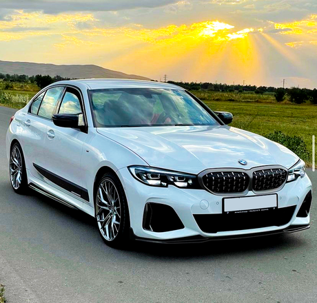 A white BMW sedan is parked on a road, with sunlight streaming through clouds in a grassy landscape. The license plate reads, "Premium Selection - 11011210 g73/11476 Omringes."