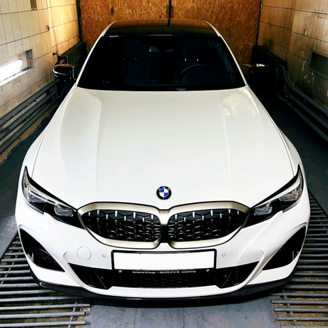 A white BMW car is stationary in an enclosed garage, with a frontal view highlighting its kidney grille. The text on the license plate reads: "Автоцентр Витаваль +996 (312) 57 77 70 www.bmkg.kg".