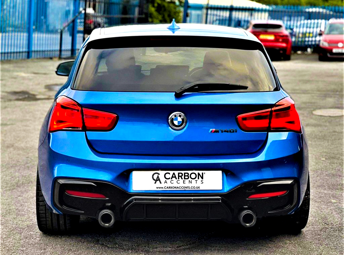 A blue BMW M140i is parked on an asphalt surface, surrounded by other vehicles. Text on the license plate reads: "CARBON ACCENTS, WWW.CARBONACCENTS.CO.UK."