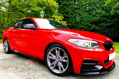 A red sports car, parked on a gravel driveway, is surrounded by lush green trees, with a classic lamp post in the background.