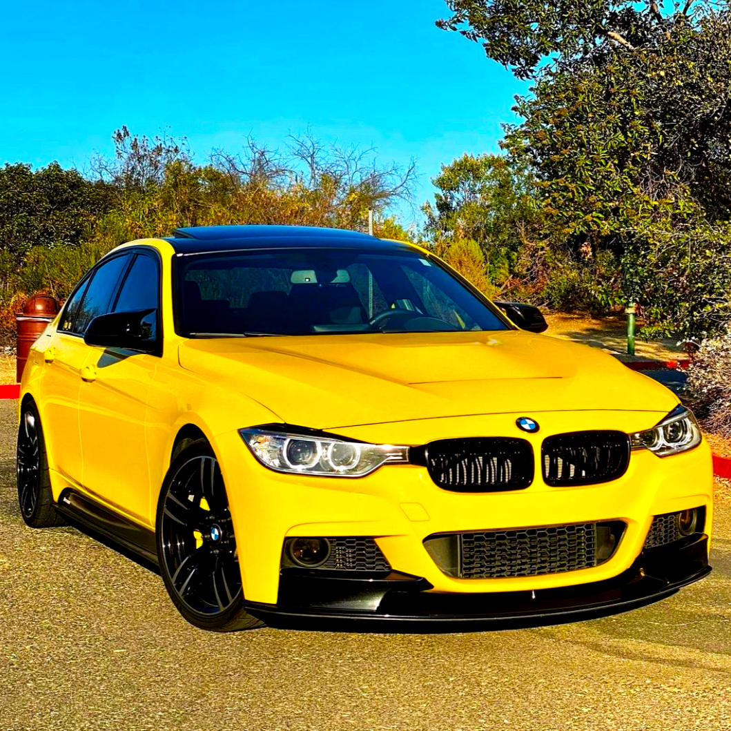 A yellow BMW car is parked, facing forward on a sunny driveway, surrounded by trees and shrubs under a clear blue sky.