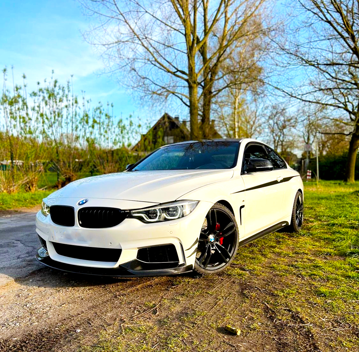A white sports car with sleek black rims is parked on dirt, surrounded by trees and greenery under a clear blue sky.