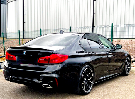 Black BMW sedan parked next to a fence, with shiny rims and distinctive red taillights, positioned on a concrete surface beside a large industrial-looking building.
